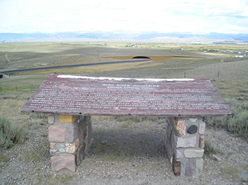 Photograph of the Trappers Point historic marker: a large, weathered sheet of painted red wood sitting across the top of two stone and mortar pedestals on the top of a hill overlooking a vast valley. The wood has words painted in white including the title: 'Wind River Mountains.'